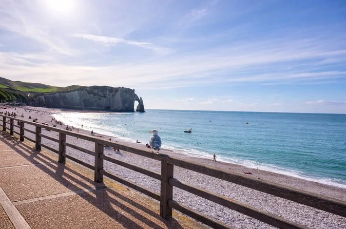Une rambarde de promenade au-dessus de la plage de galets, avec l'arche d'Aval au bout de la baie
