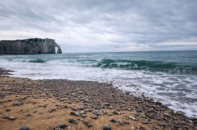 Wellen über dem Kieselstrand, im Hintergrund die Aval-Klippe und ihr Felsbogen unter bewölktem Himmel